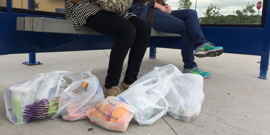An image of two people, shown from waist-down, sitting on a bench at a bus stop, with bags of groceries at their feet.