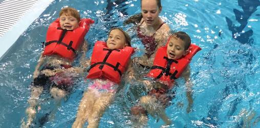 Three young children in life jackets practice floating while a teacher supports them.