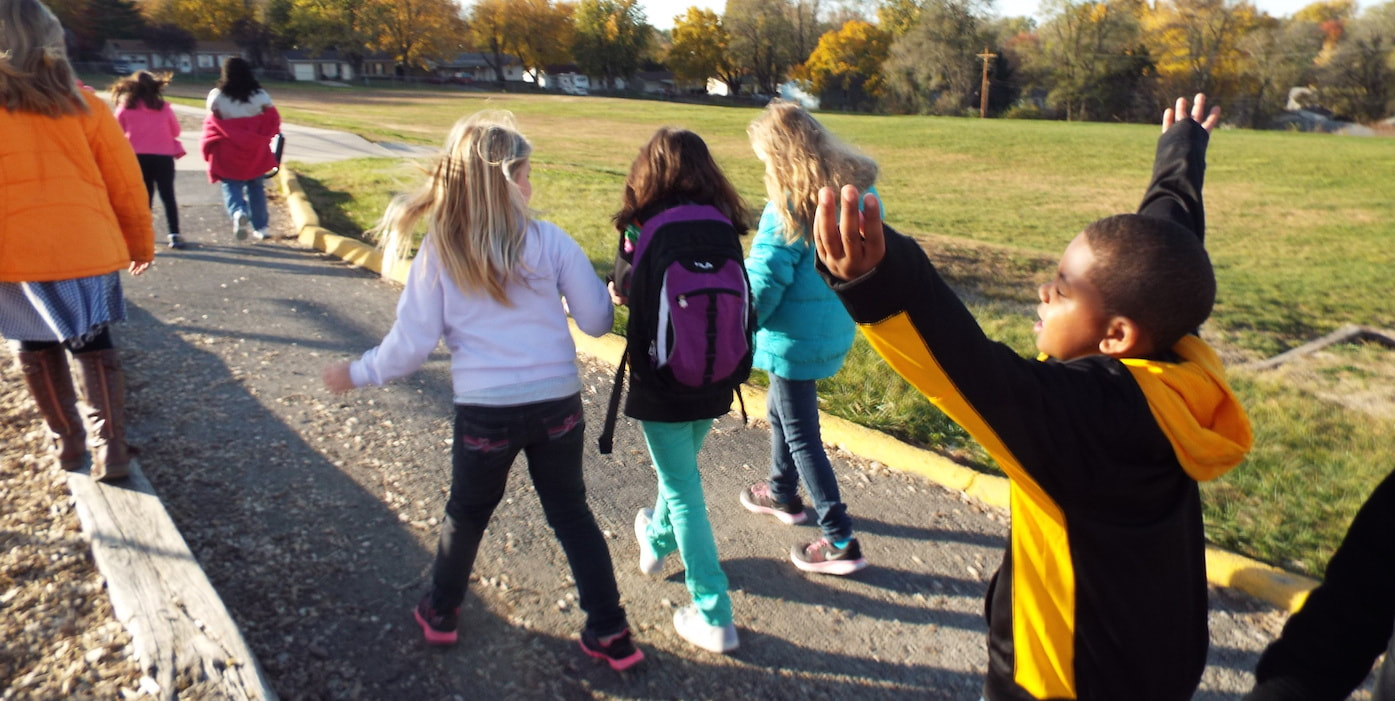 A group of elementary kids walk to school on a paved path through a park. One boy raises his arms up to  stretch them.