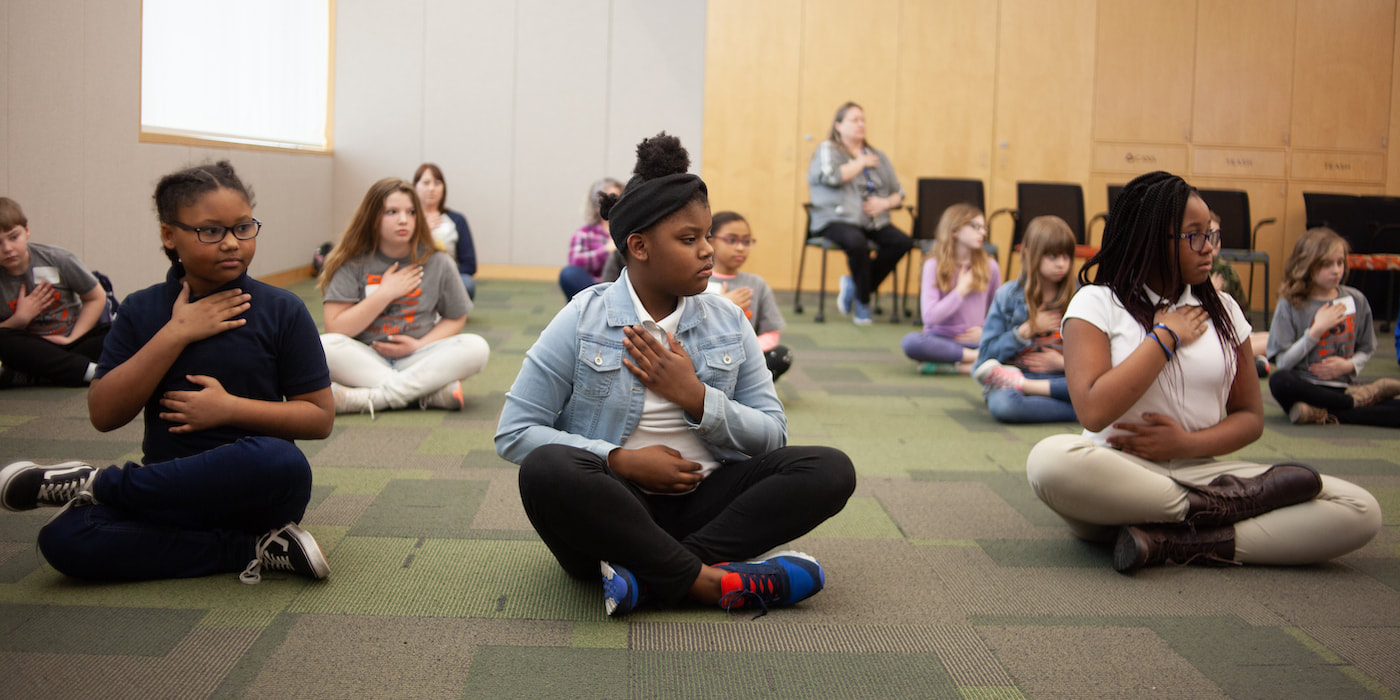 A group of middle school girls sit on the floor with their legs crossed and hands on their chests and stomach to practice mindful breathing.