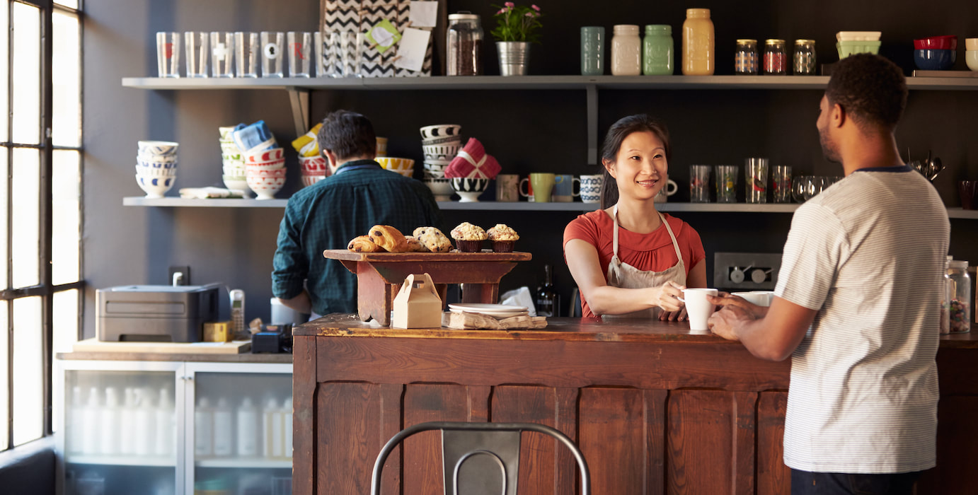 A woman serves a copy of coffee over the counter to a man while another man works with his back turned to the camera.