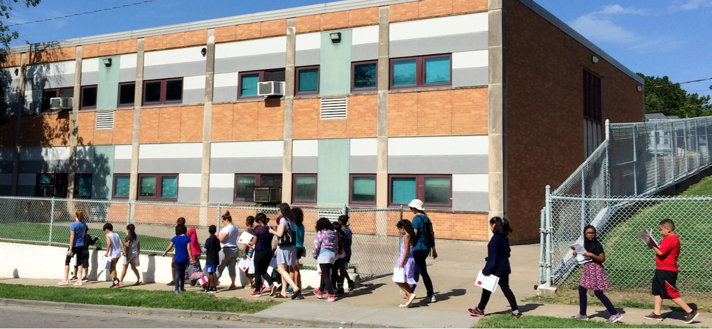 A group of kids carrying workbooks walk down a sidewalk nexct to their schoo.