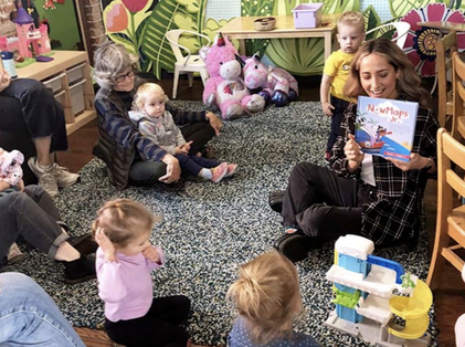 Adults and small children gather around a woman holding up a book.